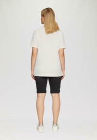 Weißes Baumwoll-T-Shirt mit kurzen Ärmeln, kombiniert mit dunklen, hochgerollten Jeansshorts und silbernen Plateausneakern. Einheitlicher Hintergrund.