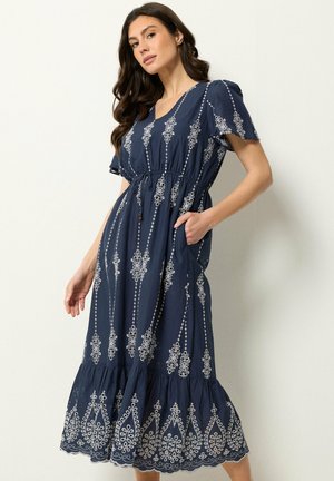 Kobieta ubrana w granatową haftowaną sukienkę midi z krótkimi rękawami, dekoltem w serek, elastyczną talią, kieszeniami i falbaną na dole, stojąca na białym tle.
