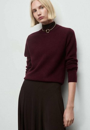 Mujer con el cabello corto y rubio, vestida con un suéter burdeos oscuro, un cuello de tortuga negro, un collar dorado tipo choker y una falda oscura y fluida, sobre un fondo liso.