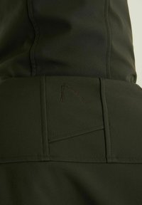 Tissu vert foncé avec une texture lisse, présentant un logo marron brodé et des coutures détaillées. Comprend des éléments de design de poche subtils.