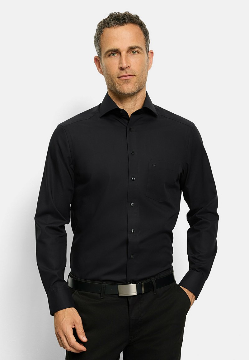 Camicia nera a maniche lunghe con bottoni, realizzata in tessuto liscio, caratterizzata da una tasca sul petto e un colletto classico. Indossata con una cintura nera.