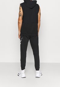 Sweat à capuche sans manches noir avec cordon de serrage, associé à un pantalon de jogging noir fuselé avec des accents blancs. Le modèle porte des baskets blanches.