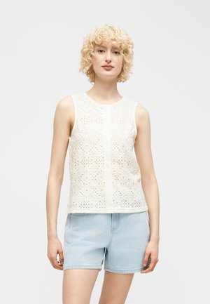 Jong persoon met kort krullend blond haar, gekleed in een mouwloos wit kanten topje en lichtblauwe denim shorts, staand tegen een effen achtergrond.