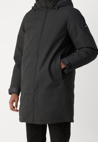 Svart isolerad parka med dragkedja framtill, huva och stora sidofickor, justerbara ärmslut och en slät, matt yta.