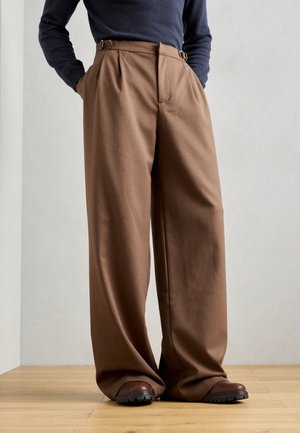 Brown, wide-leg trousers with a high waist, pleats, and belt loops, crafted from a smooth fabric. Paired with dark brown footwear.