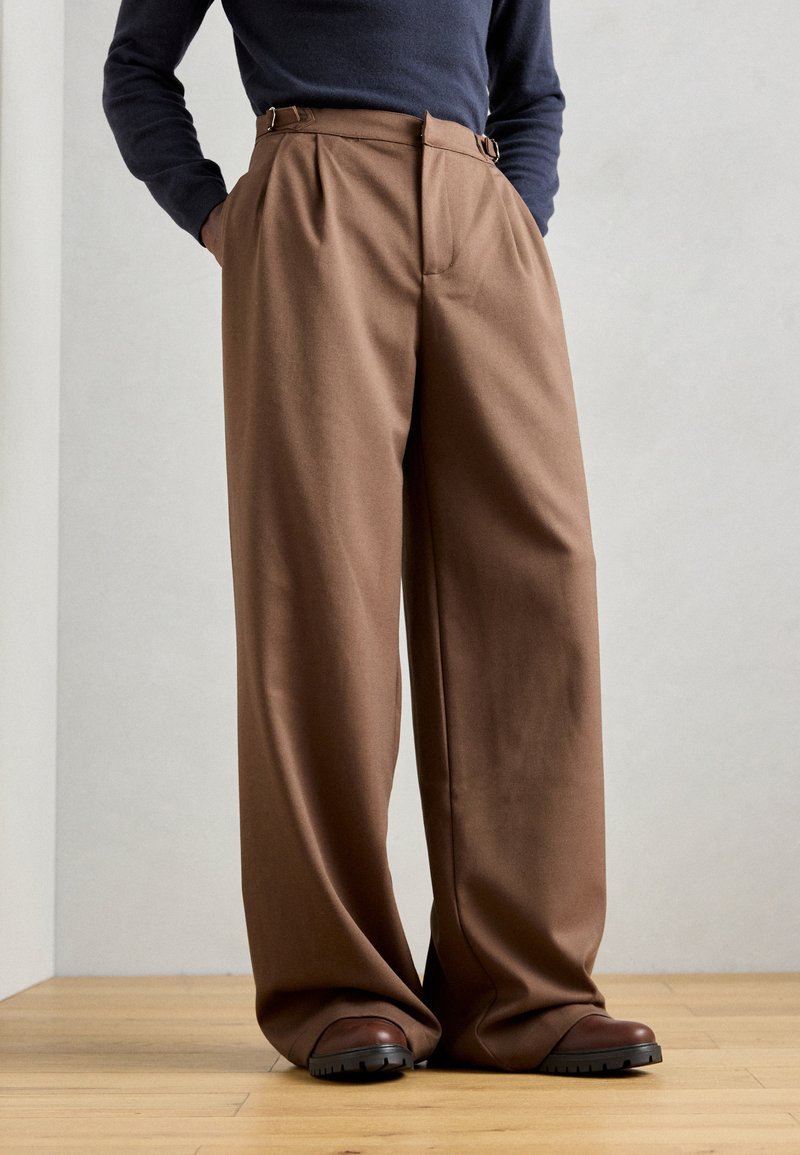 Brown, wide-leg trousers with a high waist, pleats, and belt loops, crafted from a smooth fabric. Paired with dark brown footwear.
