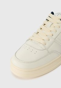 Λευκό δερμάτινο αθλητικό παπούτσι με κρέμα κορδόνια, λεία υφή και σχεδίαση με τρύπες στη μύτη. Διαθέτει στρογγυλή μύτη και διακριτικές λεπτομέρειες ραψίματος.