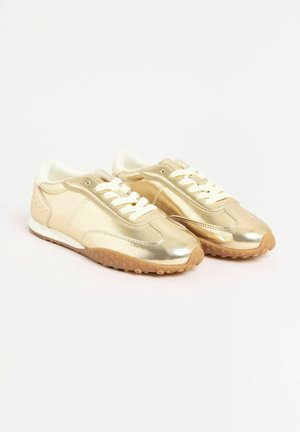 Auksiniai metaliniai sportbačiai su lygiu paviršiumi, baltais raišteliais ir guminiais padu, turinčiais taškinį raštą. Pabrėžimai šviesiai bežiniai.