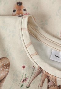 Heledat värvi kangas, millel korduv metssiga muster koos väikeste lilledega, ümara kaelusega ja pehme tekstuuriga. Silt on nähtav.