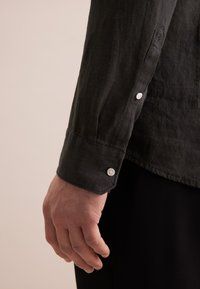 Camicia di lino nera a maniche lunghe, con polsini bottonati e una finitura testurizzata. I polsini presentano bottoni bianchi e un delicato dettaglio di cucitura.