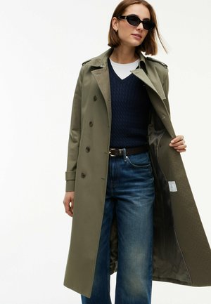Mujer con gabardina verde oliva sobre suéter azul marino, camisa blanca, vaqueros azules, cinturón negro y gafas de sol negras, sujetando la gabardina abierta.