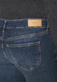 Acercamiento de la parte trasera de una persona que lleva jeans VERO MODA de color azul oscuro, con una etiqueta de cuero en la cintura y detalles de costura visibles.