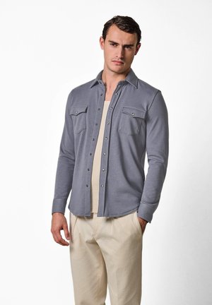 Uomo con camicia grigia chiaro abbottonata sopra canottiera beige e pantaloni beige, in piedi con una mano in tasca contro sfondo bianco.