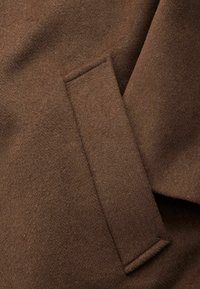 Brauner Woll-Mix-Stoff mit einer weichen, strukturierten Oberfläche, ausgestattet mit einem rechteckigen Taschendetail und einer dezenten Naht.
