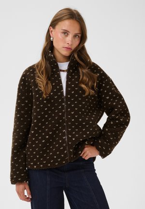 Jeune femme aux longs cheveux ondulés porte une veste polaire marron foncé à pois par-dessus un haut blanc, debout devant un fond clair uni.