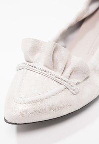 Ballerina a punta in materiale bianco lucido. Presenta un accento arricciato con una fila di piccole perline metalliche sul davanti.