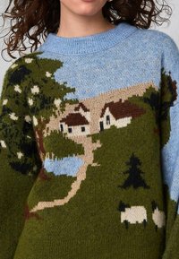 Tröja med stickad lantlig landskap som visar blå himmel, träd, två hus, en stig och två betande får på gröna kullar.