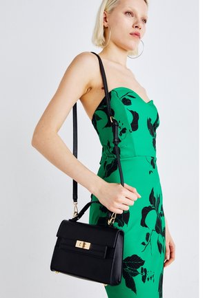 Sac à main en cuir noir avec des accessoires dorés, de forme rectangulaire, et une bandoulière amovible, tenu par une personne portant une robe verte avec des motifs floraux noirs.