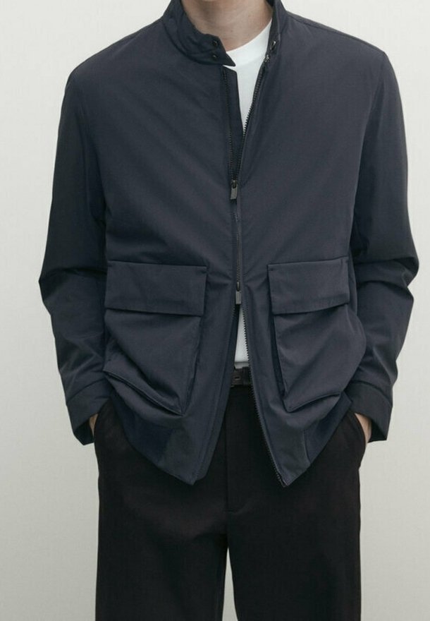 Homme portant une veste zippée bleu marine foncé avec deux grandes poches avant et un pantalon noir, mains partiellement dans les poches, sur un fond uni.