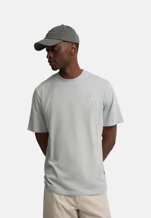 Hombre con camiseta gris claro con pequeño logo en el pecho, pantalones cortos beige y gorra de béisbol gris oscuro, mirando hacia su izquierda con las manos en la espalda.