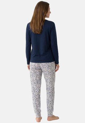 Frau steht barfuß, trägt ein dunkelblaues Langarmoberteil und weiße Pyjamahosen mit Blumenmuster, mit dem Rücken zu einem schlichten Hintergrund gewandt.