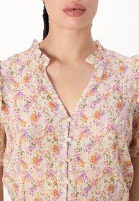 Blusa floreale di un light cream con fiori rosa, arancioni e verdi. Presenta una chiusura a metà bottone, colletto e maniche corte a sbuffo. Tessuto texturizzato.