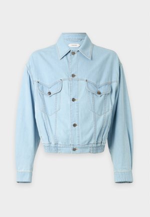 Junger Mann trägt eine hellblaue Jeansjacke, schwarzes Poloshirt mit rotem Kragen, schwarze Hose und schwarze Schuhe mit goldenen Zehenkappen, steht vor weißem Hintergrund.