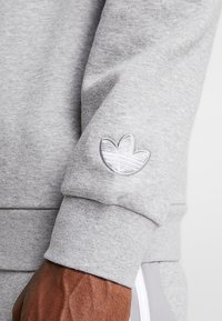 Grå sweatshirt med texturerad tyg, med en vit broderad logotyp på ärmen och ribbat manschett. Enkelt design med en figurnära form.