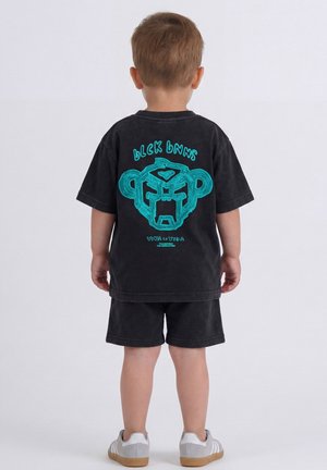 Jong kind met zwart T-shirt en korte broek met een blauw gestileerd gorillaontwerp op de rug, staat met de rug naar de camera op een effen achtergrond.