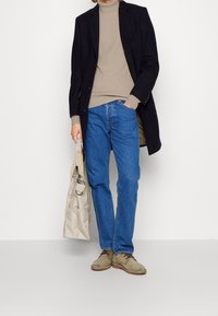 Marineblazer, beigem Ripp-Pullover, blauen Jeans und grauen Schuhen. Hält eine beigefarbene Tragetasche. Lässiges Ensemble mit unterschiedlichen Texturen und Farben.