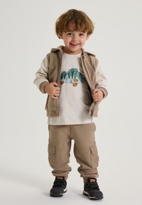 Lys beige zip-up hættetrøje over en hvid t-shirt med et fiske graphic, parret med beige cargo bukser og sorte sneakers. Blødt, afslappet materiale.