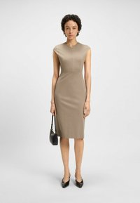 Knee-length, form-fitting dress in light brown fabric with a deep V-neck and cap sleeves. Worn with black pointed-toe heels and a small handbag.