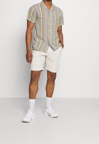 Uomo che indossa una camicia a maniche corte a righe multicolori, pantaloni corti beige, calze bianche e sneakers bianche, in piedi con le mani in tasca.