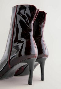 Sorte patentlæder ankelsko med høj hæl, der har røde syninger langs sømmene og en glat, blank overflade.