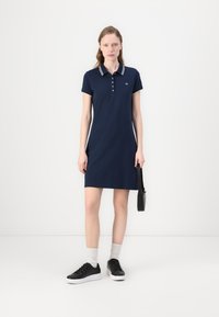 Mekko, valloittava laivastonsininen, pehmeästä puuvillasta valmistettu. Siinä on kolmen napin kaulus, valkoiset raidat, lyhyet hihat ja suora leikkaus.