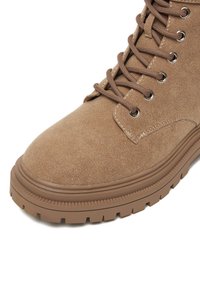 Brązowy zamszowy but z teksturowanym wykończeniem, okrągłym noskiem i wytrzymałą gumową podeszwą. Posiada metalowe oczka i płaskie sznurowadła.