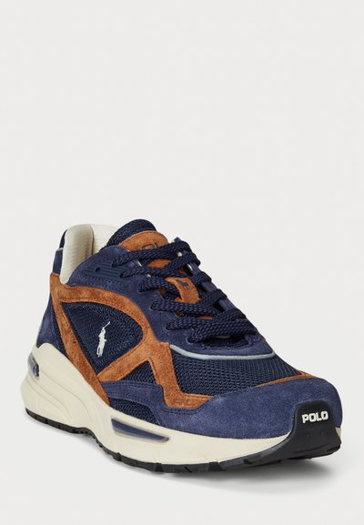 Zapatilla azul marino con acentos de gamuza marrón, paneles de malla y una suela acolchada. Presenta cordones tonales y un logo de la marca prominente en el talón.