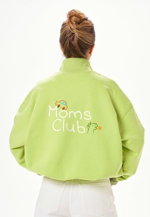 Femme aux cheveux blonds en chignon porte une veste vert clair avec le texte « Moms Club », une petite voiture et un dinosaure vert dans le dos.