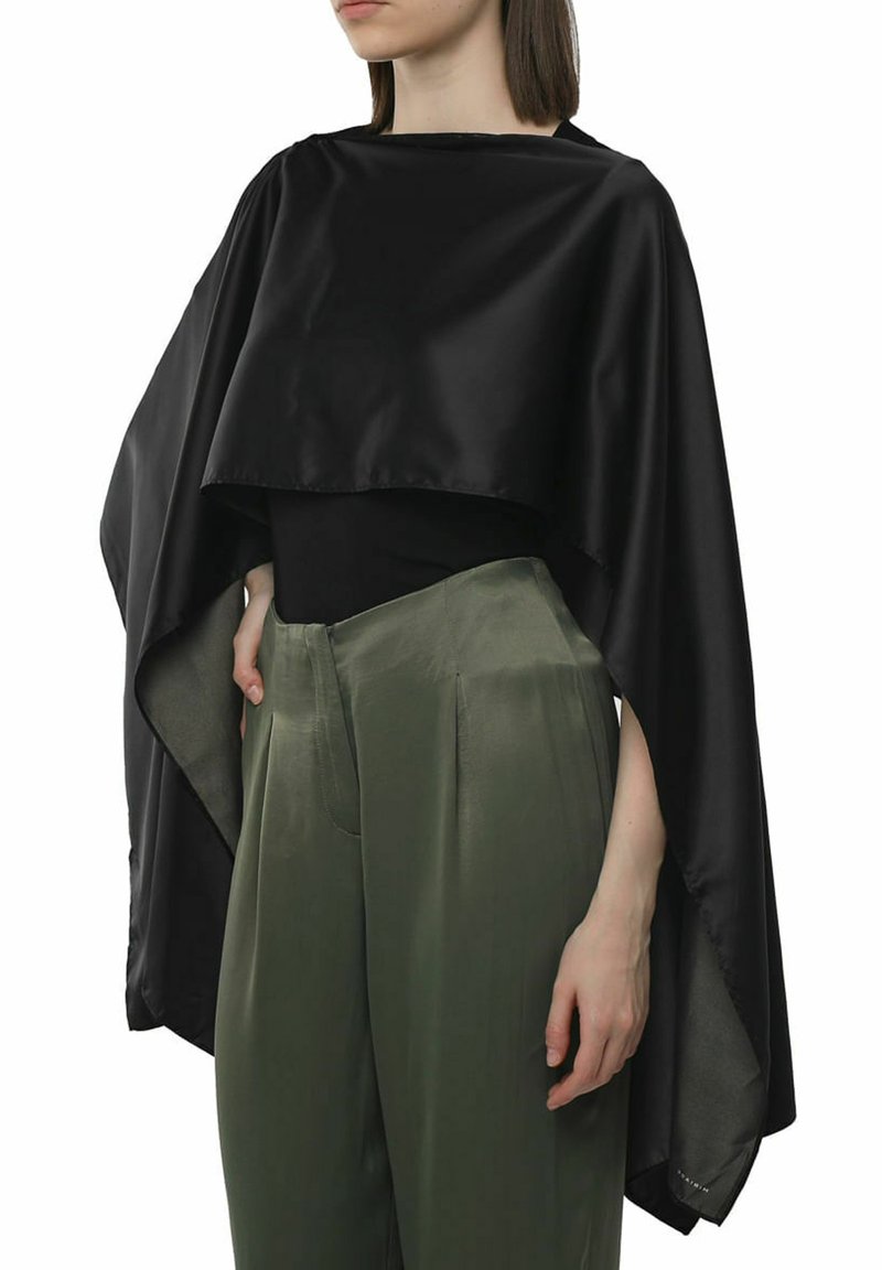 Cape cropped en satin noir avec un ourlet asymétrique, porté sur un haut noir et un pantalon taille haute vert olive. Texture douce, design léger.
