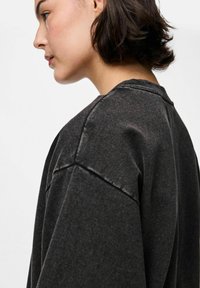 Jaunas žmogus su trumpais tamsiais plaukais, dėvintis išblukusią juodą didelį megztinį, pavaizduotas profilio pusėje prieš paprastą foną.