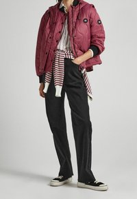 Gemusterte kastanienbraune Jacke mit Druckknöpfen, kombiniert mit einem gestreiften Hemd und dunklen, weiten Hosen. Schwarz-weiße Sneakers runden das Outfit ab.