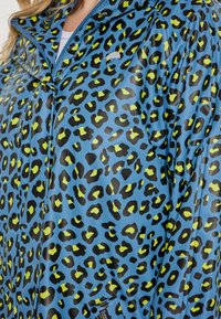 Modrá ľahká bunda s lesklým povrchom, so vzorom leopardie kože v čiernej a limetkovej zelenej. Obsahuje zipsový predný diel a zacvakávacie uzávery.