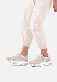 Sneakers atletici beige con tomaia testurizzata, suola bianca e un accento grigio. Presentano lacci e un logo sul lato.