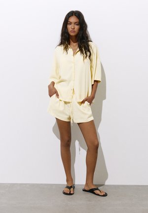 Femme aux longs cheveux foncés portant une chemise ample jaune pâle boutonnée et un short assorti, debout les mains dans les poches et des sandales noires.