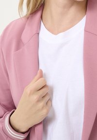 Blazer rose clair avec un col cranté, associé à un t-shirt blanc. Le blazer présente un tissu texturé et des accents rayés sur les manches.