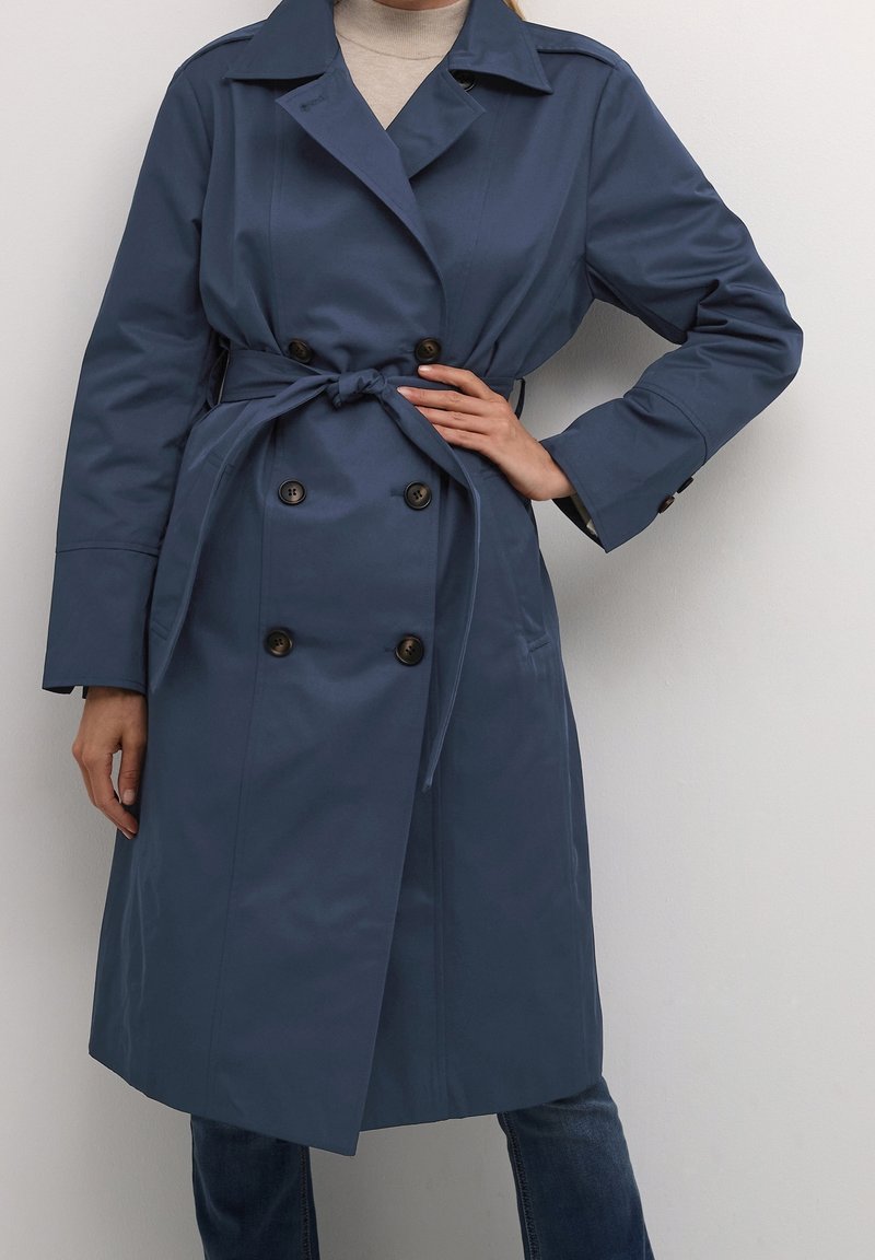 Femme portant un trench-coat bleu marine croisé noué à la taille par-dessus un col roulé beige et un jean foncé, debout devant un fond uni.