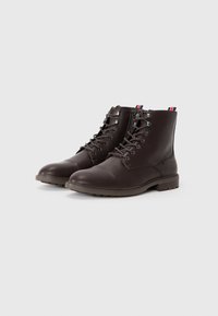 Botas de cuero marrón hasta el tobillo con punteras redondas, diseño con cordones, ojales de metal y una superficie texturizada. Presenta una lengüeta a rayas en la parte trasera.