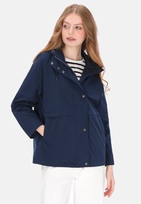 Granatowa wodoodporna kurtka z zamkiem z przodu, dwoma dużymi kieszeniami, kapturem ze sznurkiem oraz luźniejszym krojem. Pod spodem widoczna pasiasta koszula.