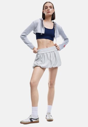 Grijze cropped hoodie, marineblauw ribbed crop top, grijze geplooide rok, witte sneakers met zwarte strepen, witte enkelsokken, gladde textuur.