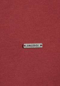 Tessuto rosso con finitura texturizzata, con un'etichetta argento attaccata che riporta la scritta "SMILODOX" in lettere incise.